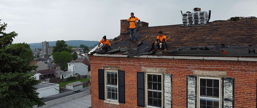 A roofing crew stands on the roof of a brick building in orange shirts, completing different types of roof damage repairs