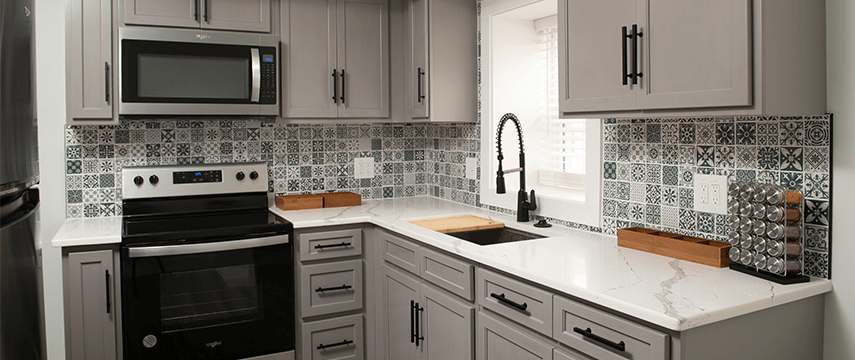 A new kitchen remodel with white counters, gray cabinets, and a gray and white tiled backsplash.