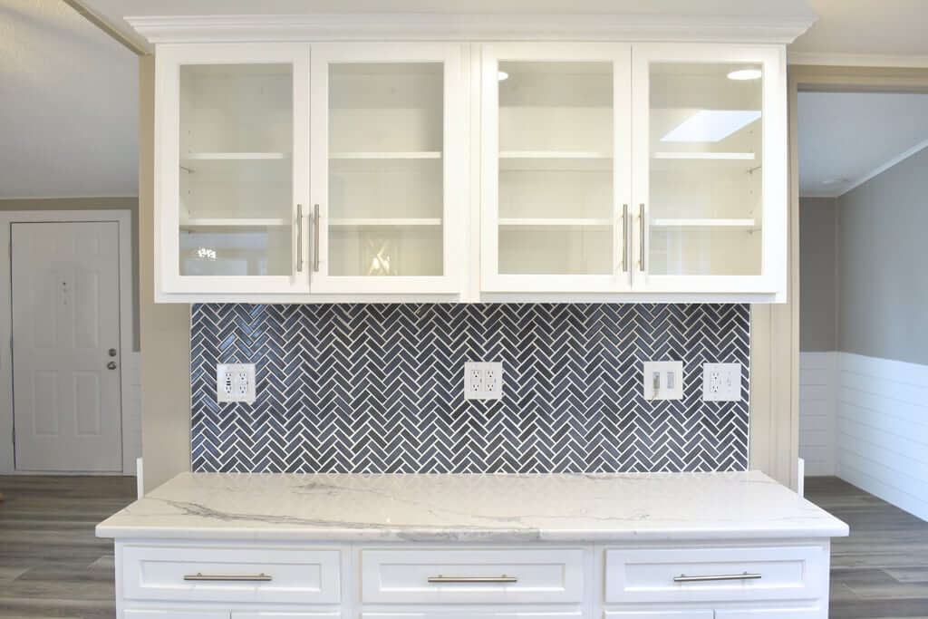 Newly remodeled kitchen showing a gray backsplash, white cabinets with glass doors, and white countertops.